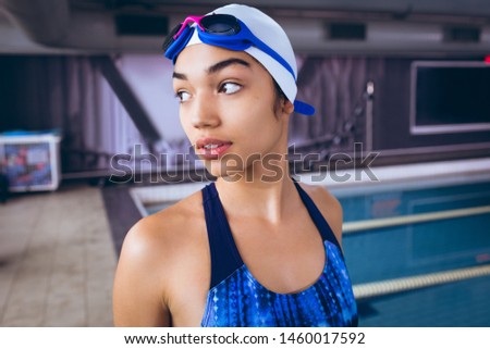 Front view of a young African-American woman wearing a swimsuit and swimming cap with goggles while standing by an olympic sized pool inside a stadium