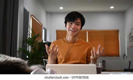 Front View Of Young Asian Man Wearing Earphones And Having Video Call With Colleagues.