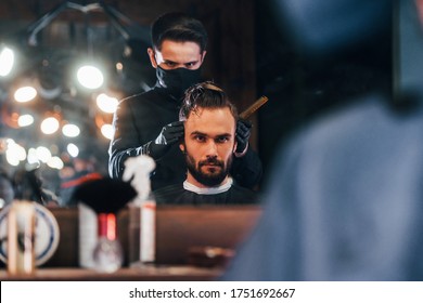 Front View Of Young Bearded Man That Sitting And Getting Haircut In Barber Shop By Guy In Black Protective Mask.