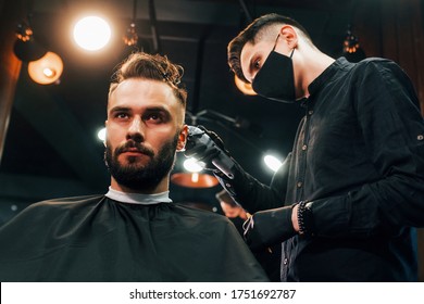 Front View Of Young Bearded Man That Sitting And Getting Haircut In Barber Shop By Guy In Black Protective Mask.