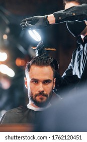 Front View Of Young Bearded Man That Sitting And Getting Haircut In Barber Shop By Guy In Black Protective Mask.