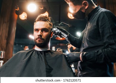 Front View Of Young Bearded Man That Sitting And Getting Haircut In Barber Shop By Guy In Black Protective Mask.