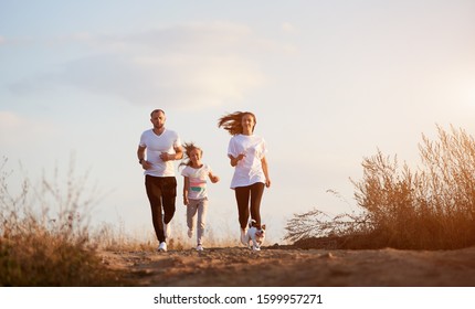 Front View Of Young And Beautiful Family Of Three Jogging With Their Dog Outside The City On The Village Road On The Setting Sun