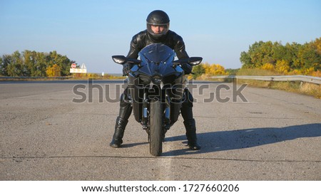 Front view of young biker in helmet and leather jacket is sitting on motorcycle at highway road and ready to ride. Man is going to ride at sport motorbike. Concept of freedom and adventure. Close up.