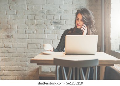 Front view of young businesswoman sitting at table in coffee shop and talking on cell phone while drinking coffee and using laptop. In background white brick wall. Freelancer working outside office.