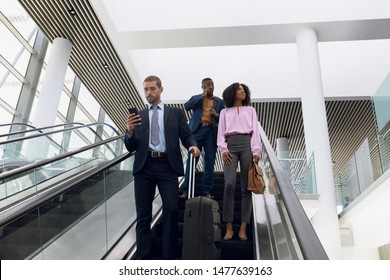 Front view of a young Caucasian businessman looking at smartphone while he stands with a suitcase on an escalator in a modern building, behind him a young African American businesswoman and a young - Powered by Shutterstock