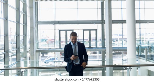 Front View Of Young Caucasian Businessman Looking Down Smiling At His Smartphone. He Is Standing Leaning On A Handrail In The Glass Walled Atrium Of A Modern Business Lobby. Modern Corporate Start Up