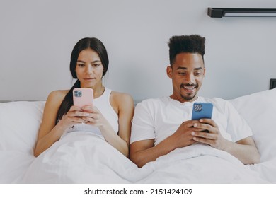 Front view young couple two family man woman in casual white clothes lying in bed hold in hands use mobile cell phone read chat text rest spend time together in bedroom lounge home in own room house