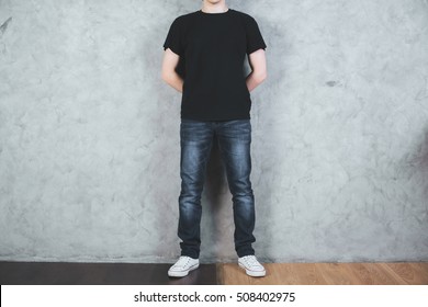 Front View Of Young Man In Empty Black Shirt And Jeans On Concrete Wall Background. Mock Up