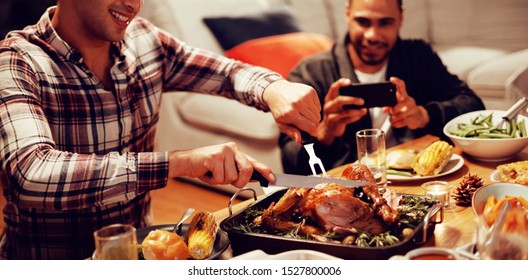 Front View Of A Young Mixed Race Man Sitting At A Table For Thanksgiving Dinner At Home With Friends Carving The Turkey, A Young Mixed Race Male Friend Taking A Photo With His Smartphone In The