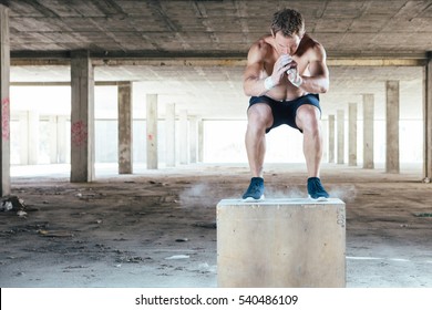 Front view of young muscular man with naked torso doing box jumps in abandoned building. 