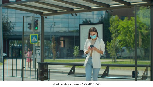 Front view of young woman using smartphone while waiting at public transport stop. Girl in medical face mask looking at phone screen while browsing internet. Health and safety, quarantine. - Powered by Shutterstock