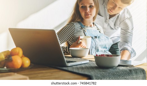 Front View.Morning, Breakfast.Young Married Couple In Kitchen.Pregnant Woman Is Sitting At Table And Using Laptop, Man Is Holding Her Pregnant Belly, Looking On Computer Monitor. Lifestyle.