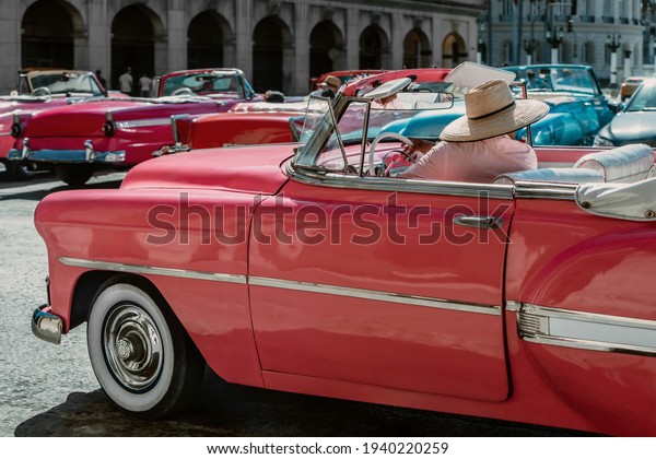 Front and wide angle photo of a light pink color old\
American car parked in the streets of Havana, Cuba. Driver sitting\
in front. Car surrounded by similar old style American cars.\
February 15, 2018