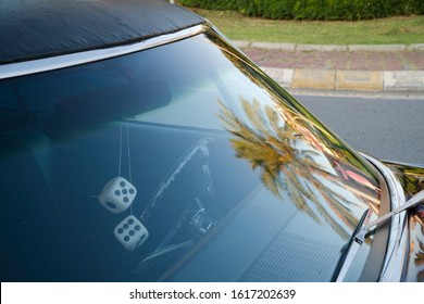 Front Window Detail Of A Purple Classic American Car At Street. There Are Dice Shaped Retro Style Car Ornament On Rear View Mirror And A Palm Tree Reflection On Windshield .