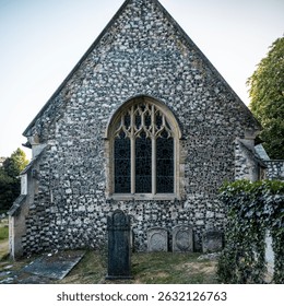 Frontal close-up of a large Gothic stained-glass window set within a flint gabled wall of an old church, with several weathered tombstones in the immediate foreground. - Powered by Shutterstock - Get 15% off with code: PIKWIZARD15