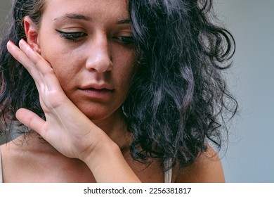 Frontal close-up portrait of young woman bringing her hand to touch the skin of her cheek and chin with the back. She may be feeling how smooth it is, but she may be sad and afraid of a slap. - Shutterstock ID 2256381817