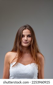 frontal half-length portrait of charming and attractive young woman with long brown hair, she wears a white sheath dress and her shoulders leave her skin bare, she smiles quietly and confidently - Shutterstock ID 2253960925