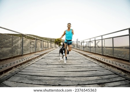 Frontal photo of an adult runner with an amputated arm running on an outdoor bridge with a dog attached to a harness. Canicross concept. Activities with a border collie. Running with pets.