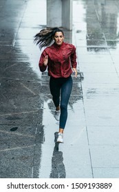 Frontal Photo Of A Sportswoman Running Wearing A Red Windbreaker, Black Leggings And White Trainers Under The Rain In The City