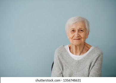 Frontal portrait of smiling elderly grey haired woman seating on a chair, wearing comfortable grey V-type sweater at home. Glasses off.