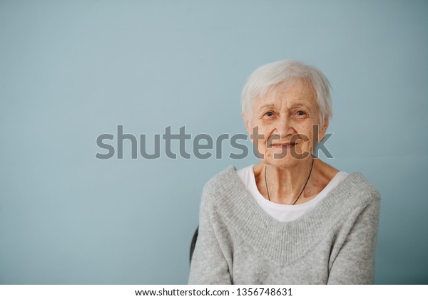 Frontal portrait of smiling elderly grey haired\
woman seating on a chair, wearing comfortable grey V-type sweater\
at home. Glasses off.