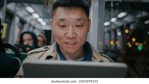 Frontal view of Chinese man traveling in public transport. Going home after work in evening. Light flickering in window outside. Male using tablet device or laptop. Actively searching for information. - Powered by Shutterstock