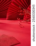 A frontal view of a crimson surface with an empty space for presentation products, surrounded by red envelopes, a winterberry branch, and some red paper fans, perfect for the Asian New Year theme.