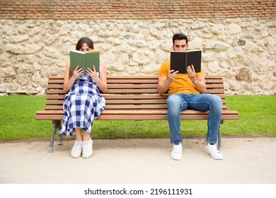 Frontal View Of Two People Sitting On A Bench Of A Park Reading Books