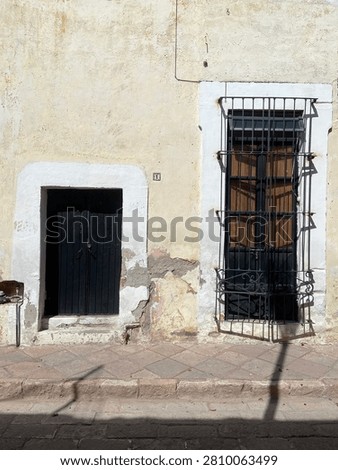 Frontal view of a weathered, rustic cream wall featuring a low black wooden door and a tall shuttered window with black iron security bars.