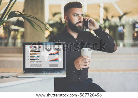 Fronte view. In foreground is laptop with graphs, charts and diagrams on screen. In background young bearded businessman sitting and talking on cellphone while drinking coffee. Online education.