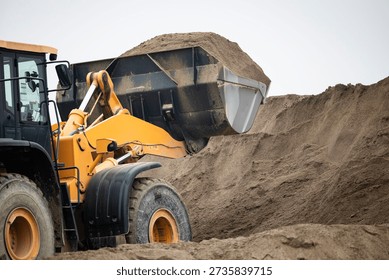 Front-end loader working at a construction site or quarry, actively scooping a large bucketful of sand from a substantial pile, highlighting earthmoving and heavy equipment operations