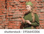A front-line nurse in the Red Army uniform from World War II mourns against a red brick wall.