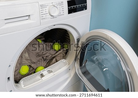 A front-loading dryer is filled with a puffy down jacket and bright tennis balls, demonstrating a laundry method to prevent clumping and ensure even drying for down-filled garments.