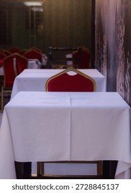 Front-view of an empty restaurant interior featuring neatly aligned tables covered with white tablecloths and red upholstered chairs. The shallow depth of field and soft warm lighting create a calm, cozy mood inside a narrow dining space. Ideal for concepts related to hospitality, fine dining, cafe ambience, event preparation, restaurant reservations, or quiet dining experiences with no people present.
