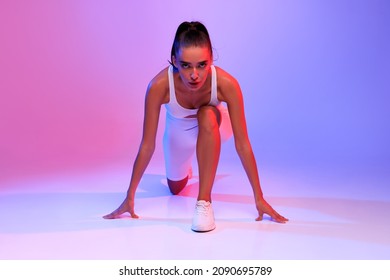 Front-View Of Motivated Female Runner Doing Crouch Start Looking At Camera Ready For Race Posing On Pink And Blue Neon Studio Background. Fit Woman Preparing For Running Workout