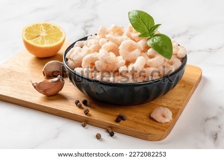Frosen shrimps in a bowl prepared for cooking. Boiled peeled prawn tails, lemon, garlic and pepper on a cutting board. Seafood recipe concept. Cooking low calorie healthy meal. Front view.