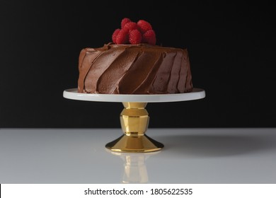 Frosted Chocolate Cake With Raspberries On A Gold Cake Stand With A Black Background And White Countertop.  