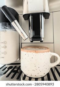 Frothed milk flows from the spout of an automatic coffee machine into a speckled ceramic mug filled with espresso. Creamy cappuccino preparation captured up close at home.