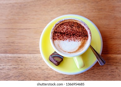 Frothy Cappuccino Coffee In A Yellow Cup And Saucer With A Hand Crafted Chocolate On The Side