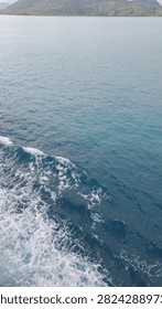 Frothy white waves churn behind a moving ship, with a green island visible on the horizon