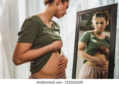 Frown young woman standing in front of a mirror and holding hands on her bloating stomach. - Shutterstock ID 2152261335