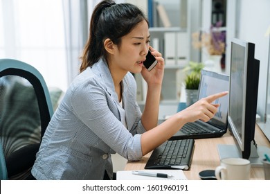 frowning asian japanese woman employee working on laptop computer at office while talking on mobile phone in light workplace. serious young girl discussing project on cellphone online point monitor