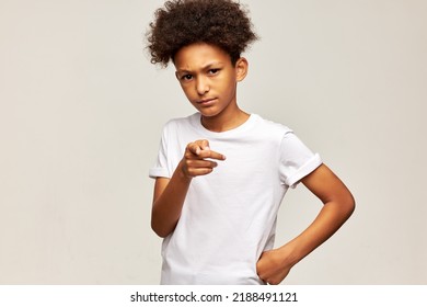 Frowning Black Teen Boy In White Mockup T-shirt With Afro Hairstyle Posing On Gray Background With Hand On Waist Pointing At Camera With Angry Facial Expression, Blaming You For Everything, Frowning