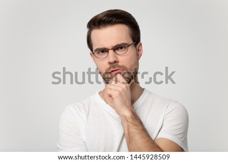 Frowning man wearing glasses white t-shirt holding hand on chin looking at camera feels distrustful studio head shot portrait isolated on grey background, concept of distrust chary suspicious person