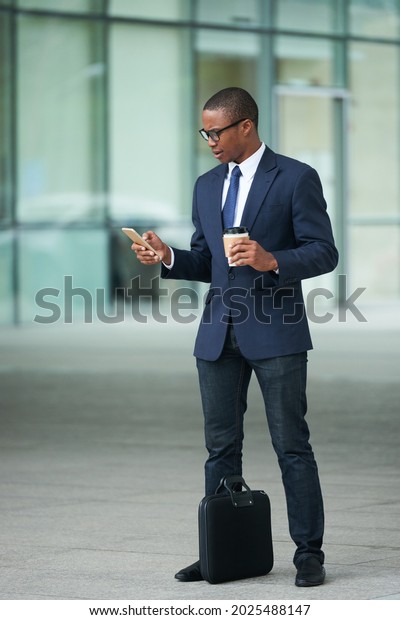 Frowning serious businessman with briefcase standing\
outside airport terminal, drinking take-out coffee and ordering\
taxi car