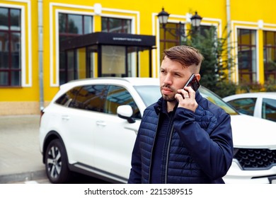 Frowning Tense Sullen Or Sad Concentrated Young Caucasian 34 Years Old Man With Goatee Beard Talks On Smartphone On Street Near White Car. Waist Up Lifestyle Business Portrait, Difficult Conversation