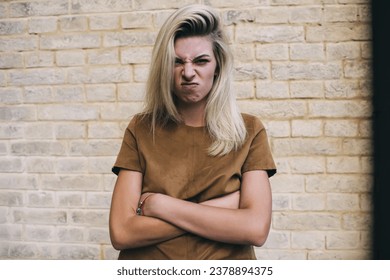 Frowning young female wearing brown shirt and standing alone with folded hands while looking at camera against brick wall in daylight and making face while waiting for friend - Shutterstock ID 2378894375