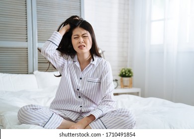 Frowning Young Girl Feeling Tired Doesn't Want Wake Up In The Morning. Unhappy Lady With Hand Scratching Hair With Itch Head. Exhausted Asian Japanese Woman Mad Sitting On Bed In Bright Bedroom.