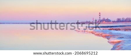 Frozen Baltic sea shore at sunset. Ice fragments and stones close-up. Lighthouse in the background. Colorful pink sky.Reflections on the water. Natural mirror. Latvia. Symbol of hope and peace